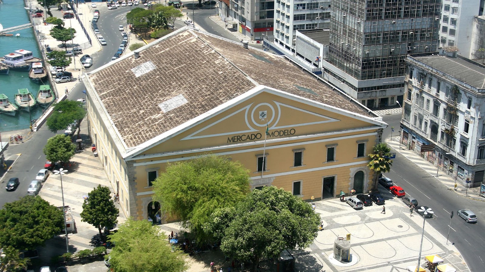 Arquitetura em Salvador Uma vista aérea ou superior do Mercado Modelo em Salvador, Bahia. O edifício principal, de fachada amarelada com detalhes brancos e um telhado de telhas marrom, está centralizado na imagem. A entrada frontal exibe o nome "MERCADO MODELO". Ao redor, há ruas movimentadas com carros estacionados e em trânsito, algumas árvores e praças. À esquerda, pode-se ver parte do porto com barcos atracados em águas azuis, e ao fundo, edifícios urbanos altos completam a paisagem.