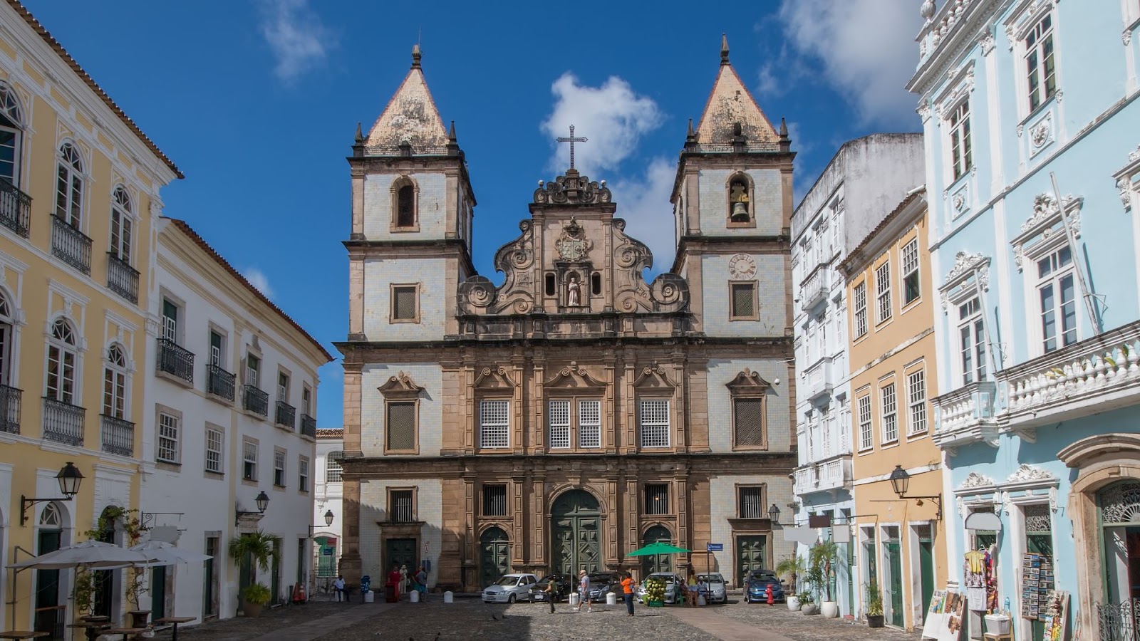 Arquitetura em Salvador Uma vista frontal da Igreja e Convento de São Francisco em Salvador, Bahia, sob um céu azul com algumas nuvens brancas. A imponente fachada barroca da igreja, em tons de bege e branco, apresenta duas torres campanárias ornamentadas com pináculos e esculturas. No centro, há um portal ricamente decorado. A igreja está situada em uma praça de paralelepípedos, onde se veem algumas pessoas e carros. Nas laterais da praça, há edifícios históricos coloridos, em tons de amarelo pálido, branco e azul claro, com janelas e sacadas.