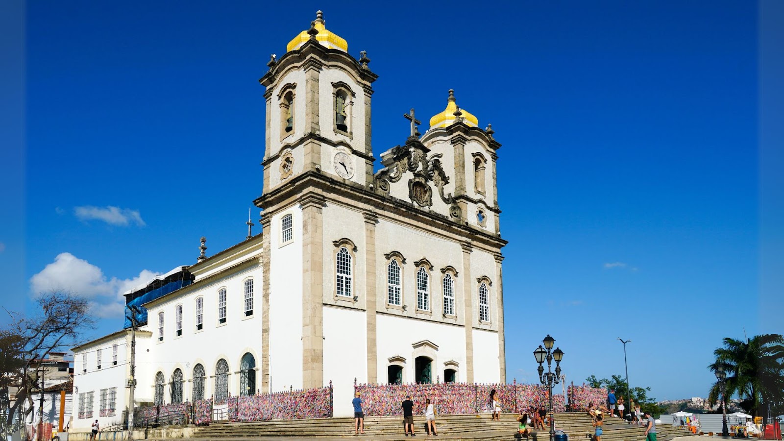 Arquitetura em Salvador Uma vista externa da Igreja de Nosso Senhor do Bonfim em Salvador, Bahia, sob um céu azul intenso e sem nuvens. A igreja branca e imponente, com detalhes em pedra escura e dourado, apresenta duas torres com cúpulas douradas e um grande relógio em uma delas. A fachada principal possui arcos e janelas características. Em frente à igreja, uma grade está coberta com inúmeras fitinhas coloridas do Bonfim, símbolo de fé e desejos. Há pessoas caminhando na escadaria e nos arredores da igreja, e algumas árvores e postes de iluminação são visíveis na paisagem.