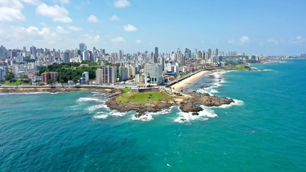 Vista aérea de uma cidade costeira com arranha-céus, praias e costões rochosos sob um céu azul brilhante.