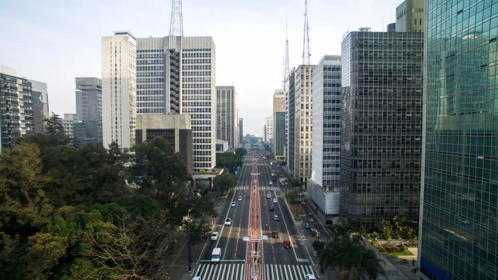 Uma ampla rua da cidade ladeada por altos edifícios modernos, com pouco tráfego e muito verde ao longo da rua sob um céu claro.