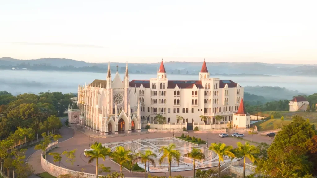 Um grande edifício branco, semelhante a um castelo, com torres vermelhas, ergue-se em meio a árvores e colinas enevoadas, banhado pela suave luz da manhã.
