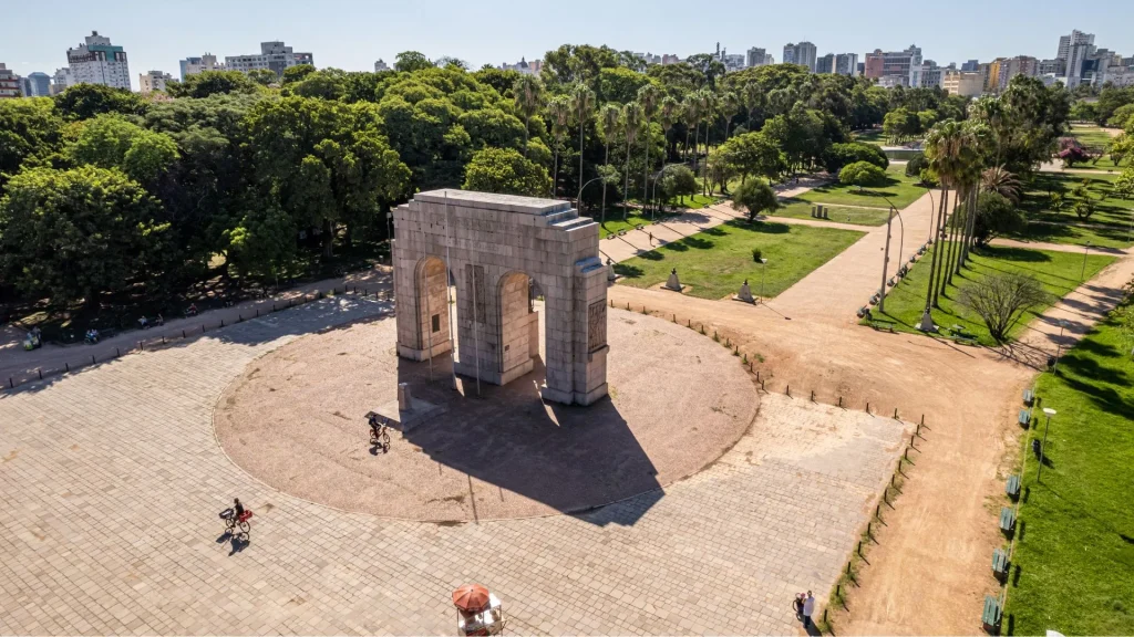 Vista aérea do Monumento à Expedicionário em Porto Alegre, localizado no Parque Farroupilha (Redenção). O monumento é um grande arco duplo de pedra clara situado no centro de uma praça circular pavimentada. Ao redor, estendem-se amplos gramados, caminhos de terra e muitas árvores altas, incluindo palmeiras. Ao fundo, é possível ver a linha do horizonte da cidade com diversos prédios sob um céu limpo. No primeiro plano, duas pessoas andam de bicicleta pela praça.
