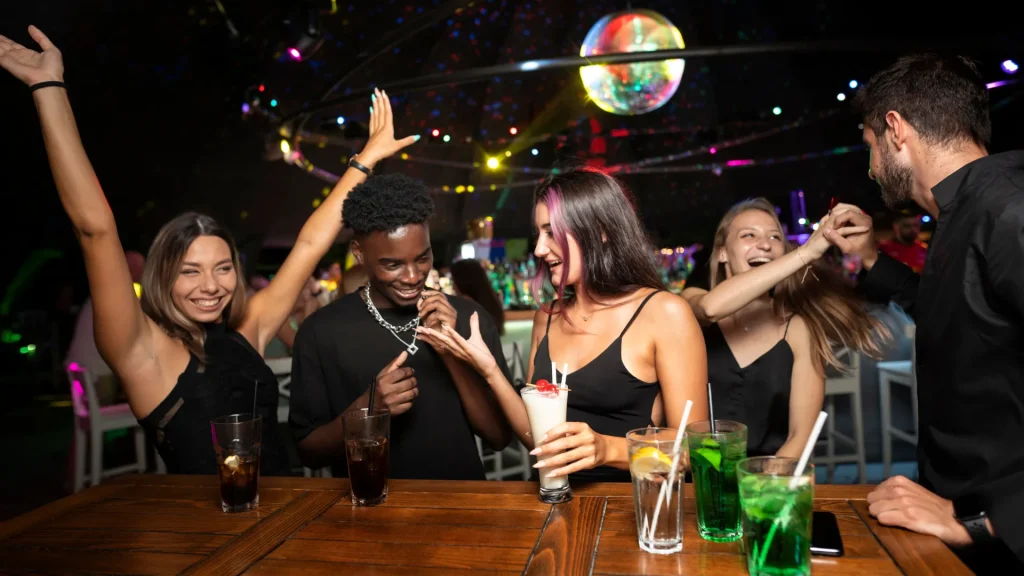 Um grupo de jovens, incluindo homens e mulheres, está rindo e socializando em um bar ou balada. Eles estão reunidos em torno de um balcão de madeira, com bebidas e copos. Ao fundo, luzes de neon e uma bola de espelhos (disco ball) pendurada no teto criam uma atmosfera de festa.