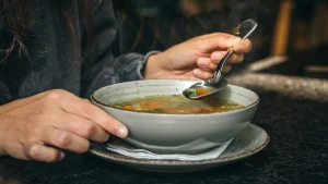 Uma pessoa segura uma tigela de sopa em um balcão ou mesa. Uma das mãos segura a borda da tigela, enquanto a outra usa uma colher para pegar um pouco da sopa, que contém vegetais.