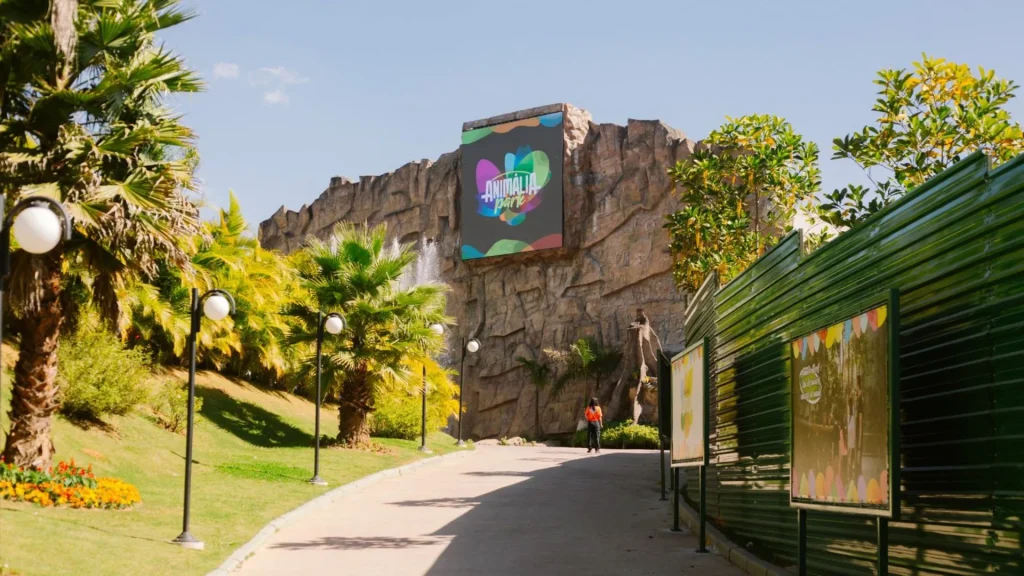 Vista de um caminho pavimentado no Animalia Park. À esquerda, um gramado com palmeiras, flores alaranjadas e postes de luz globulares. Ao fundo, uma grande estrutura rochosa artificial com uma tela digital exibindo o logotipo colorido do parque. À direita, tapumes verdes delimitam uma área em obras com cartazes informativos. O dia está ensolarado com céu azul claro.