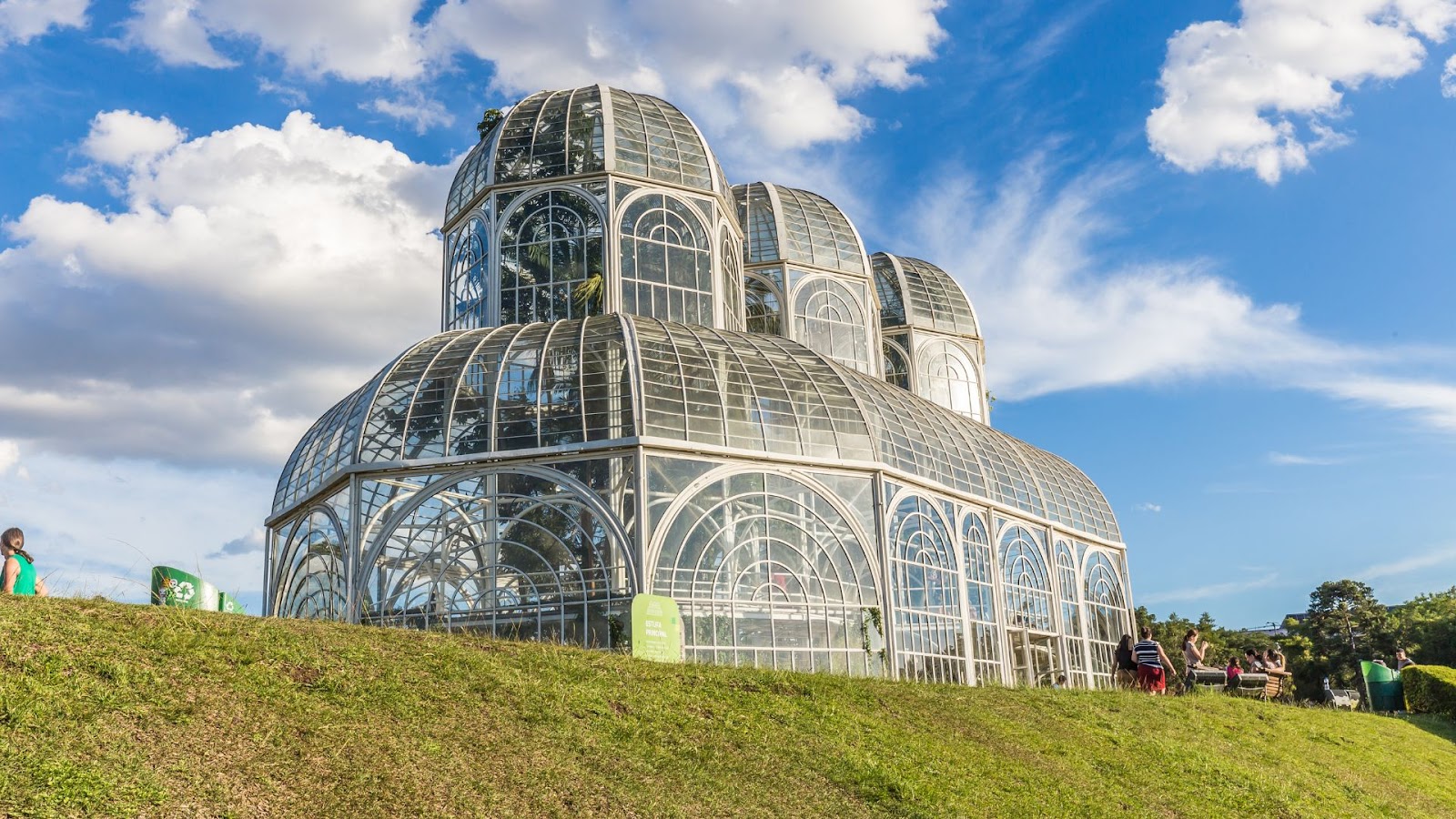 I Wanna Be Tour Jardim Botânico de Curitiba, com sua estufa de vidro em estilo Art Nouveau no centro, rodeada por gramados verdes sob um céu azul com nuvens.