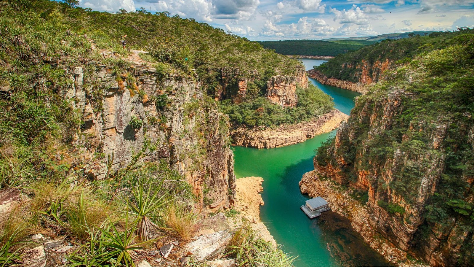 Férias de julho Vista do alto dos Cânions de Furnas, em Capitólio (MG). A imagem mostra o rio de águas verde-esmeralda correndo entre paredões rochosos e íngremes, com vegetação de cerrado no topo, sob um céu azul com nuvens.