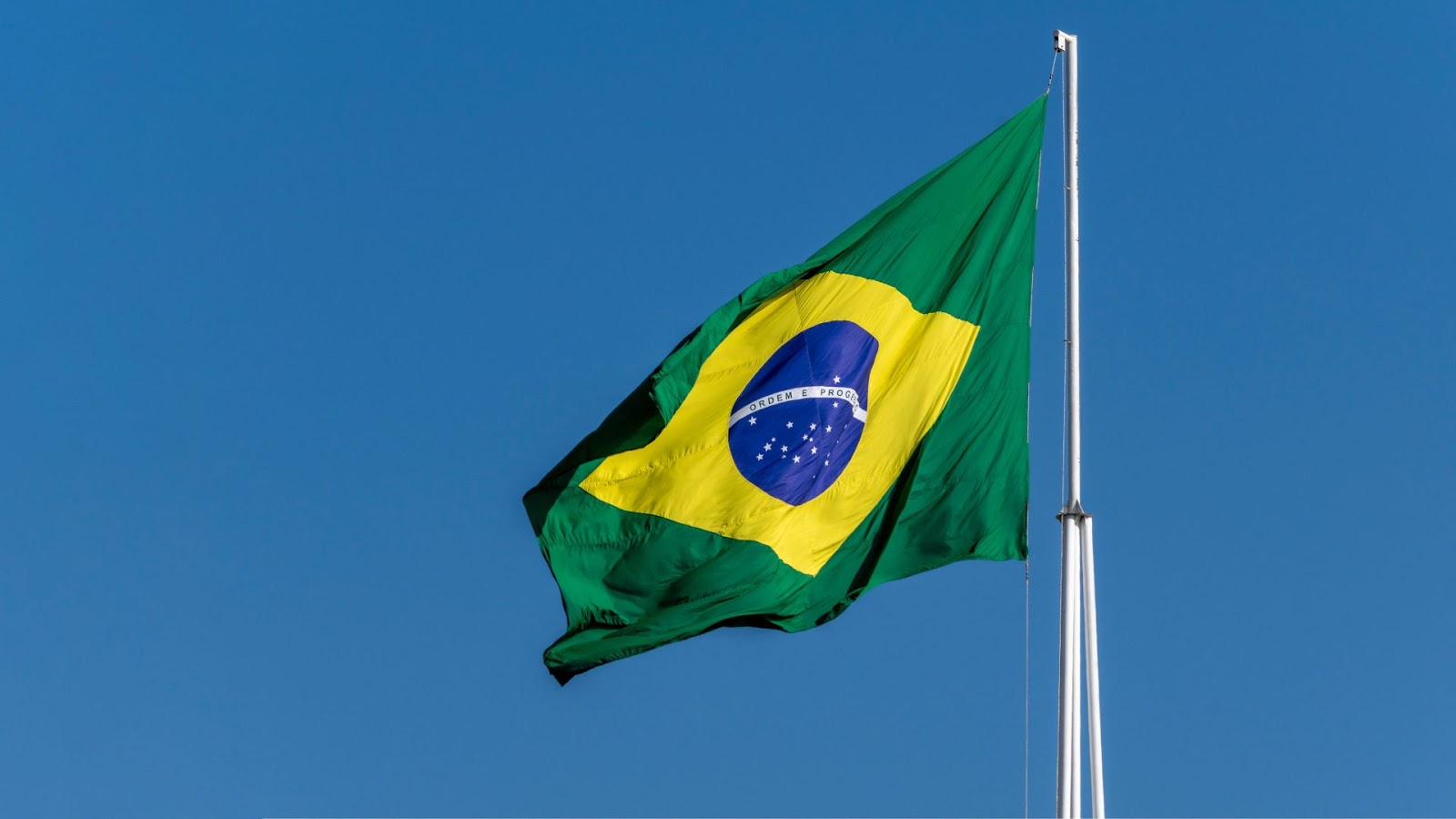 Bandeira brasileira ondulante com um losango verde e amarelo, com um globo azul e estrelas brancas, contra um céu azul claro.
