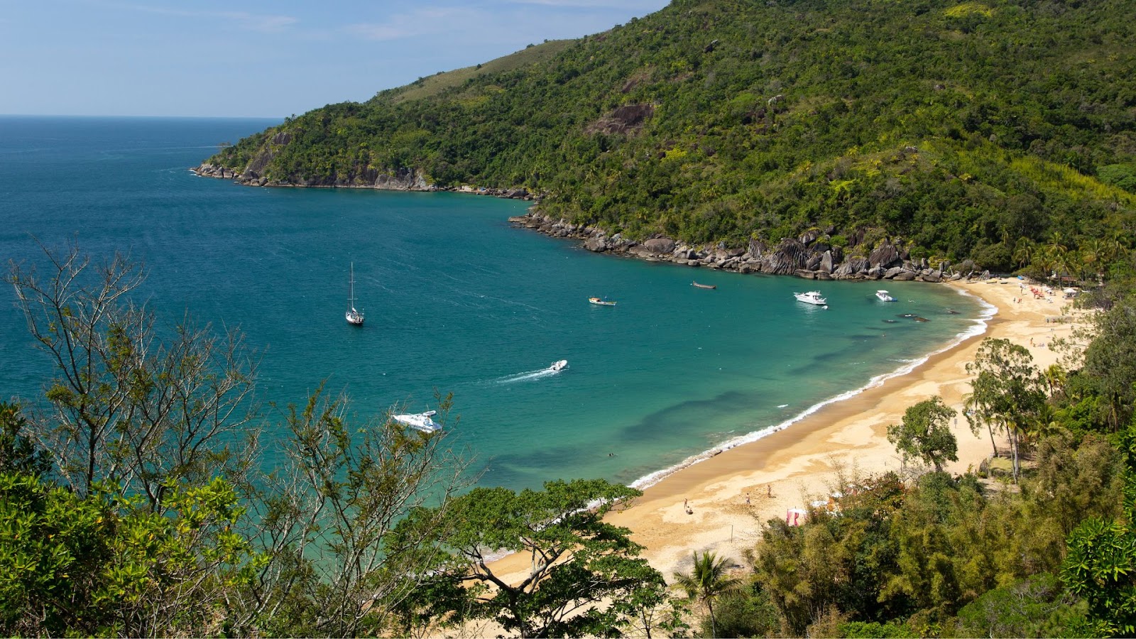Uma praia serena com areia dourada e águas azul-turquesa, cercada por colinas verdejantes e pontilhada de barcos em um dia ensolarado.
