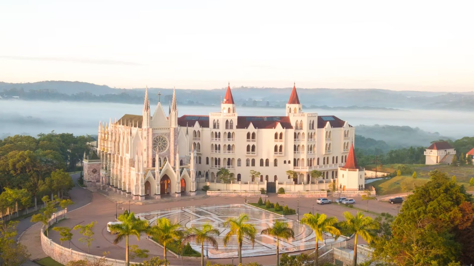 Um grande edifício branco, semelhante a um castelo, com torres vermelhas, ergue-se em meio a árvores e colinas enevoadas, banhado pela suave luz da manhã.