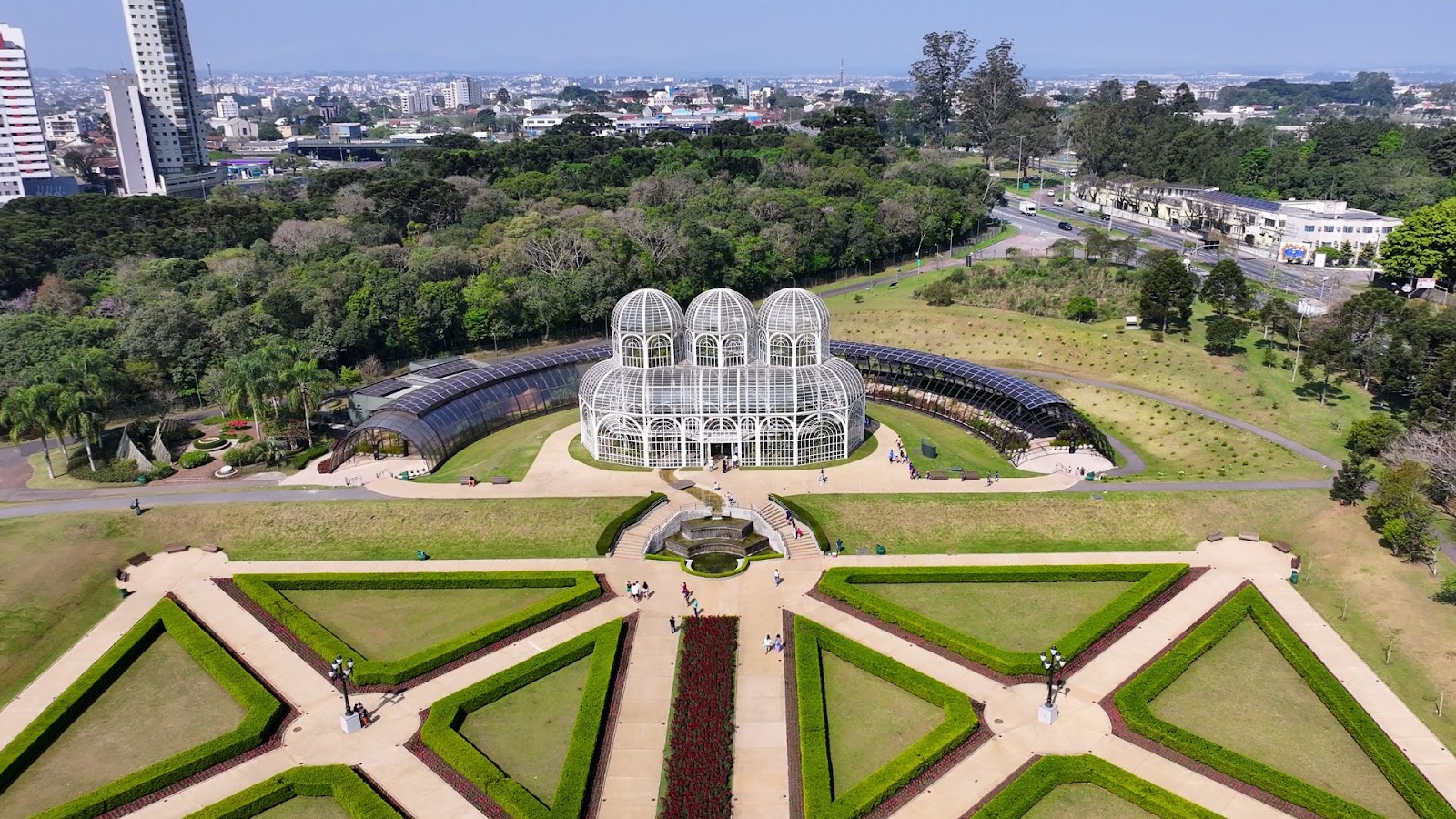 Vista aérea de um jardim botânico com uma grande estufa de vidro, sebes bem cuidadas e visitantes aproveitando um dia ensolarado.