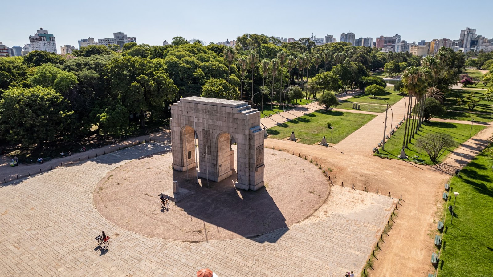 Vista aérea de um monumento em arco de pedra cercado por vegetação exuberante e edifícios urbanos, com pessoas andando de bicicleta nas trilhas.