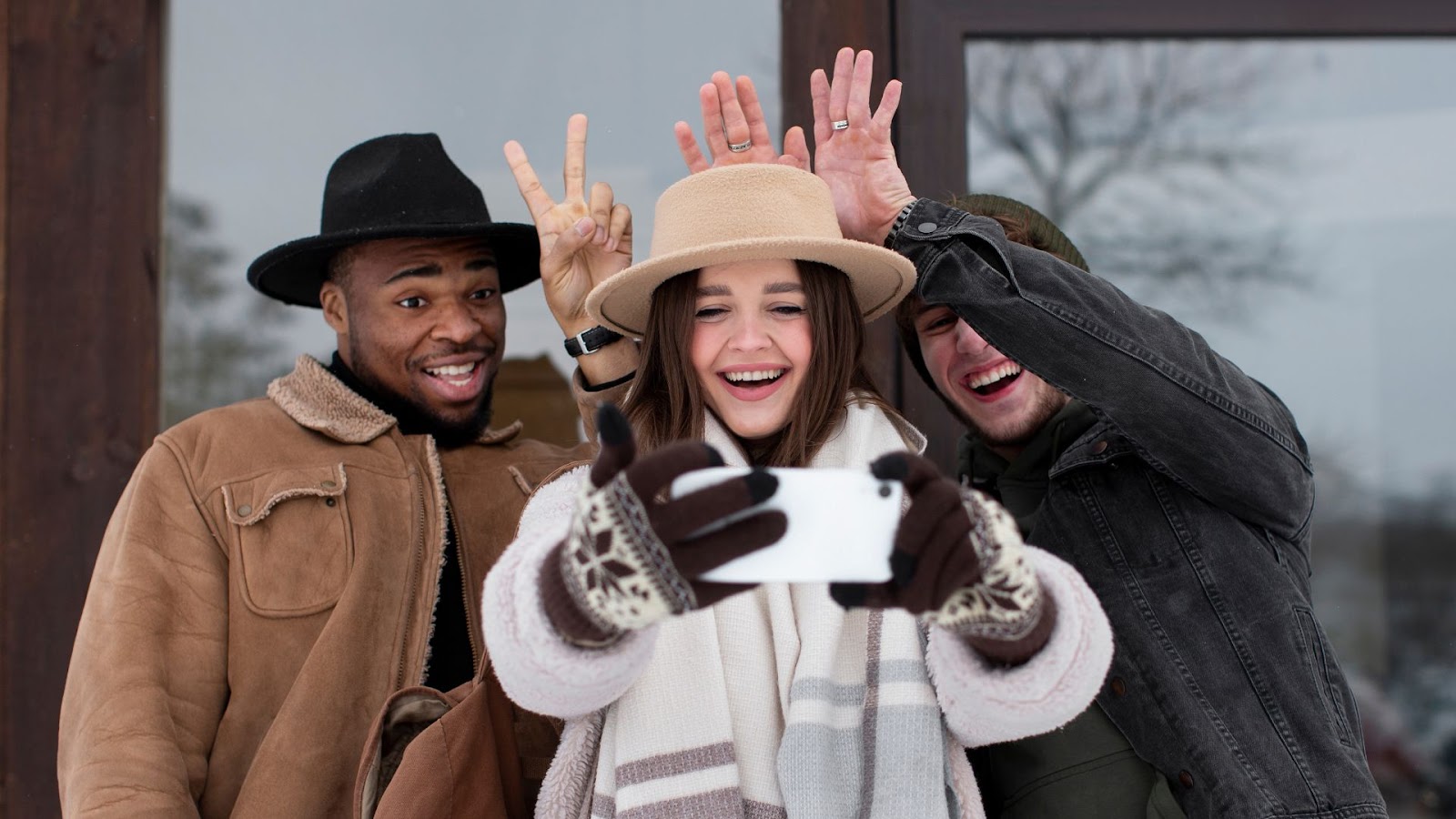 Três amigos, um homens e duas mulheres, sorriem e fazem poses engraçadas para uma selfie. A mulher no centro segura o smartphone e usa um chapéu de cor clara, luvas estampadas e um cachecol. O homem à esquerda usa um chapéu preto, e a mulher à direita faz gestos com a mão atrás da cabeça dela. Todos estão vestindo roupas de inverno.
