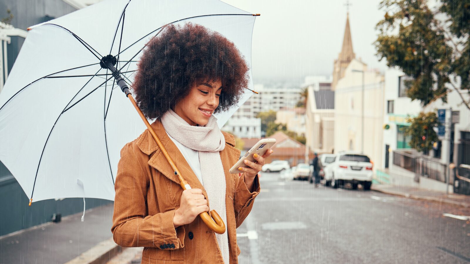 Uma mulher sorridente com cabelo afro está em uma rua da cidade em um dia de chuva. Ela segura um guarda-chuva branco e olha para a tela de seu smartphone. Ela veste uma jaqueta marrom e um cachecol claro. Ao fundo, uma igreja e outros edifícios são visíveis.