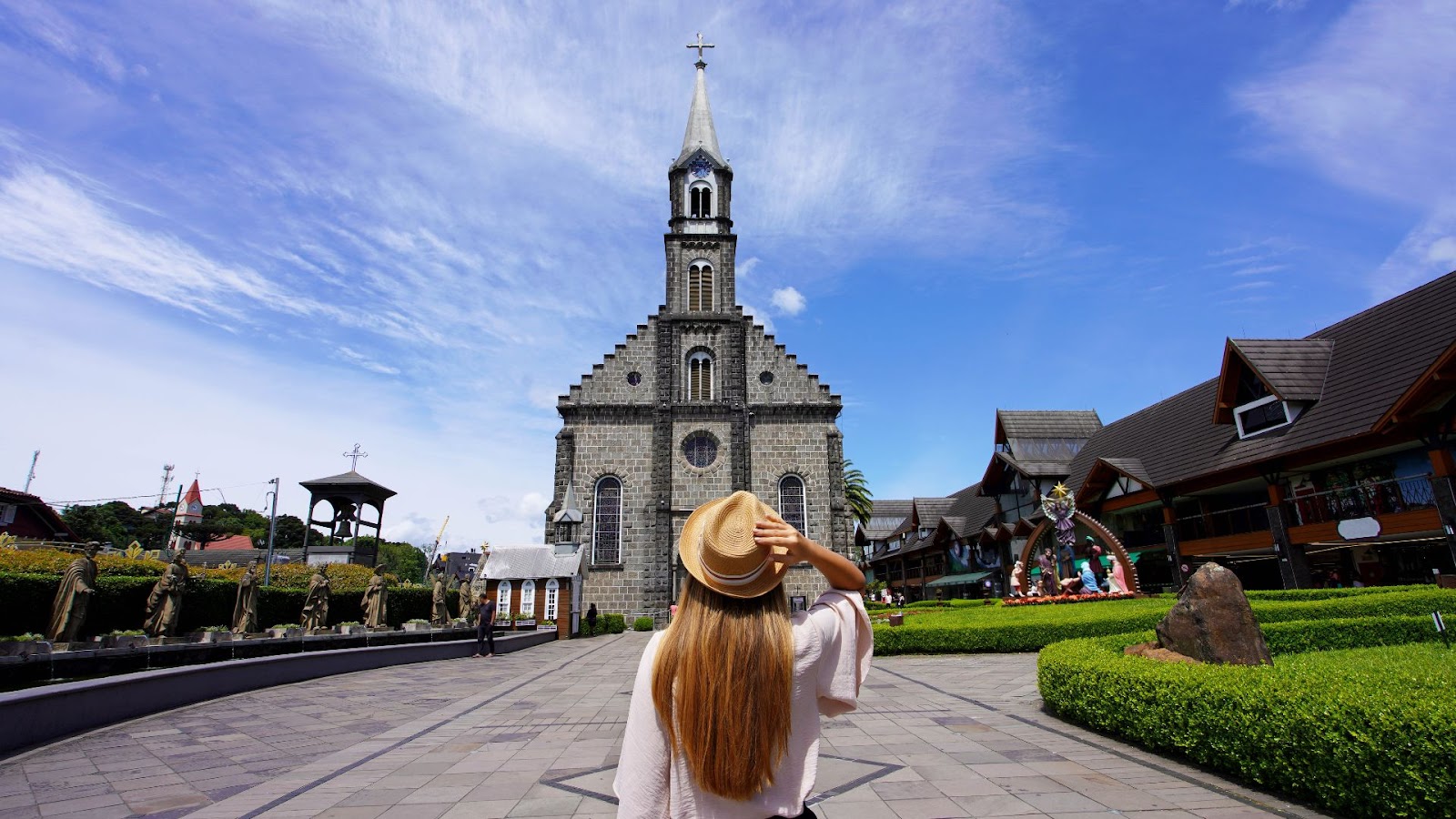 Férias de julho Uma pessoa com cabelos longos e claros é vista de costas, segurando seu chapéu de palha. Ela está no centro de uma ampla praça de pedra que leva a uma imponente igreja em estilo gótico, construída com pedras escuras. Ao lado da praça, há prédios com arquitetura de estilo europeu e jardins bem cuidados. A foto é tirada de um ângulo baixo, realçando o céu azul e vasto.