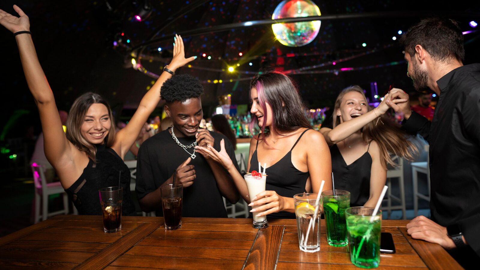 Um grupo de jovens, incluindo homens e mulheres, está rindo e socializando em um bar ou balada. Eles estão reunidos em torno de um balcão de madeira, com bebidas e copos. Ao fundo, luzes de neon e uma bola de espelhos (disco ball) pendurada no teto criam uma atmosfera de festa.