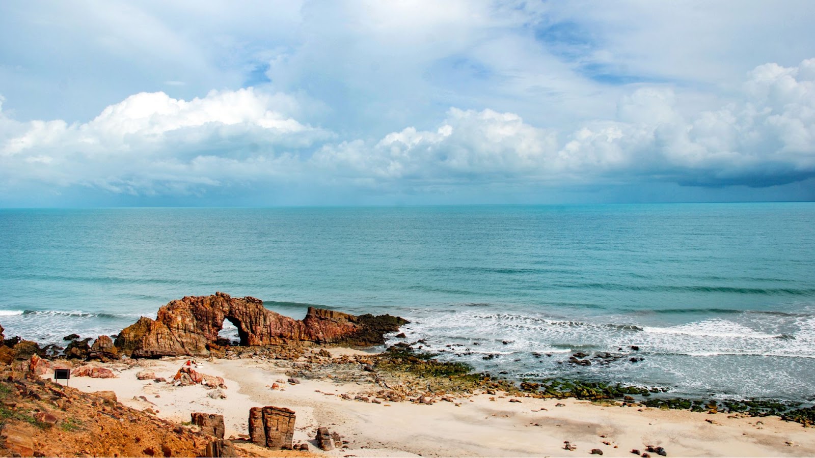 Férias de julho A famosa Pedra Furada em Jericoacoara, Ceará. A imagem mostra a grande formação rochosa de cor avermelhada com seu arco característico na beira de uma praia, com o mar ao fundo e um céu com muitas nuvens brancas e cinzentas.