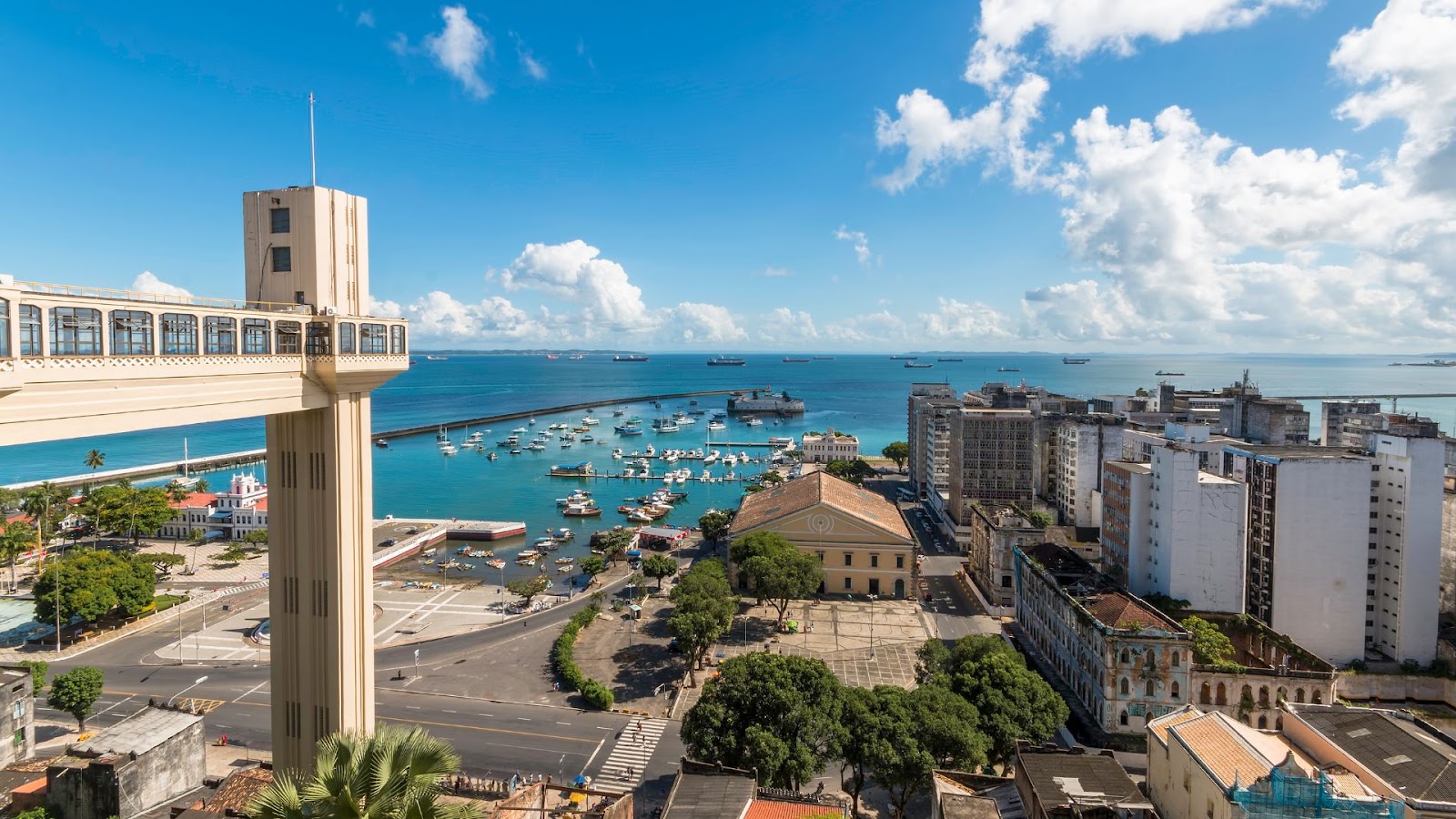 Férias de julho Vista clássica de Salvador, Bahia, destacando o Elevador Lacerda em primeiro plano à esquerda. A imagem mostra a Baía de Todos-os-Santos repleta de barcos e a Cidade Baixa, sob um céu azul ensolarado com nuvens brancas.