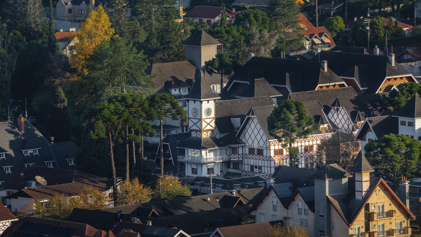 Férias de julho Uma paisagem urbana com forte influência germânica, vista de um ângulo elevado. No centro, um grande edifício branco com estrutura de madeira escura e uma torre com relógio se destaca. Ao redor, diversos telhados íngremes se misturam com a vegetação densa, que inclui araucárias e algumas árvores com cores de outono.