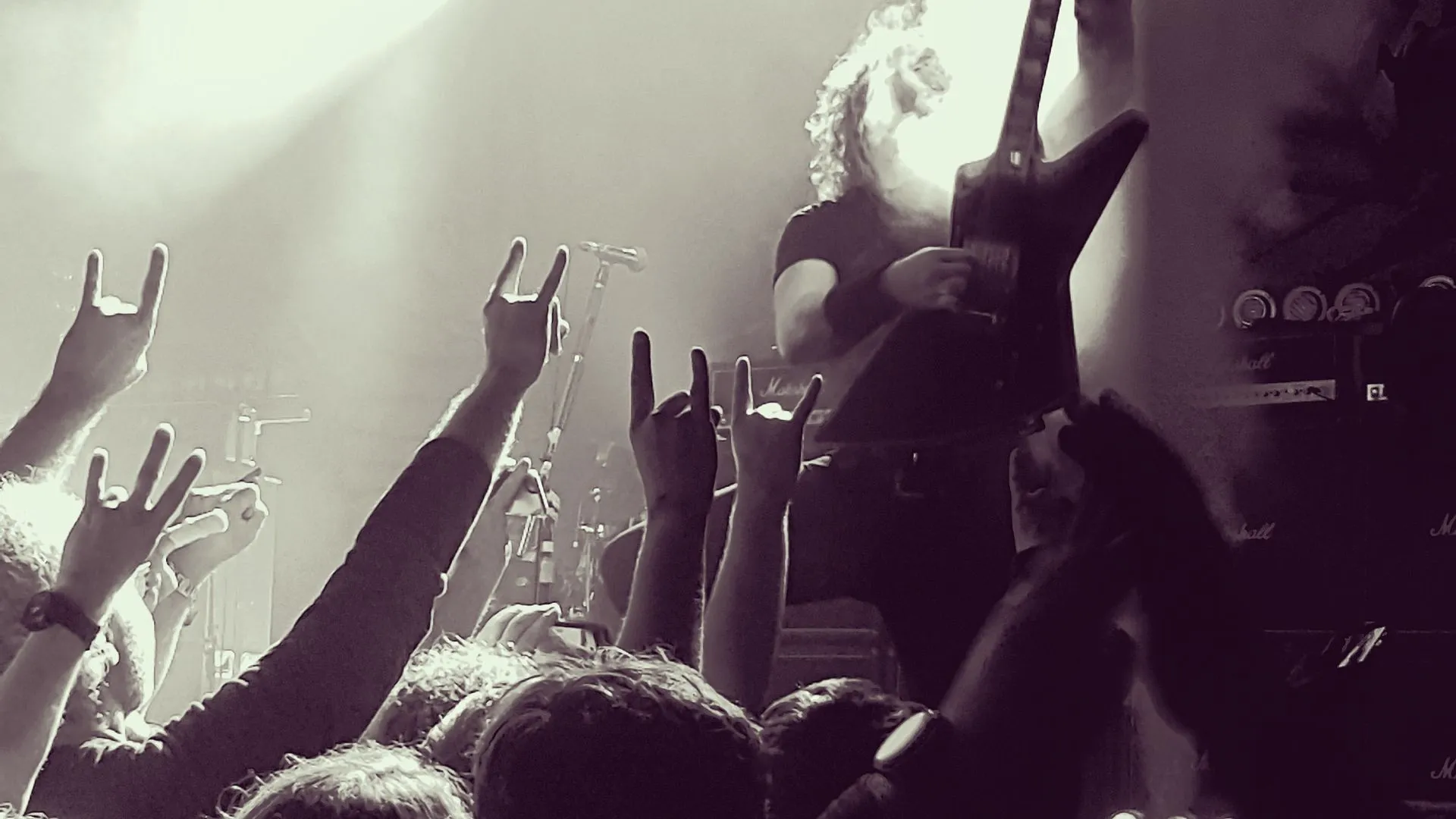Multidão em um show de rock com os braços levantados, fazendo o gesto 'chifres do rock'. No centro, um guitarrista toca, iluminado por um holofote. A imagem é em preto e branco e a atmosfera é de energia e entusiasmo.