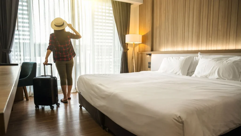 Uma pessoa com chapéu e uma mala de rodinhas está em pé em um quarto de hotel, olhando pela janela. A cama king-size, com lençóis e travesseiros brancos, ocupa grande parte do quarto. A luz do sol entra pela janela e a atmosfera é de tranquilidade e relaxamento. A decoração do quarto é moderna e elegante.