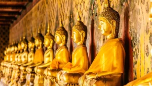 Fila de estátuas de Buda douradas e sentadas, uma ao lado da outra, dentro de um templo. As estátuas têm a mão direita pousada sobre a perna direita, na pose de meditação. A parede atrás delas é decorada com pinturas antigas.