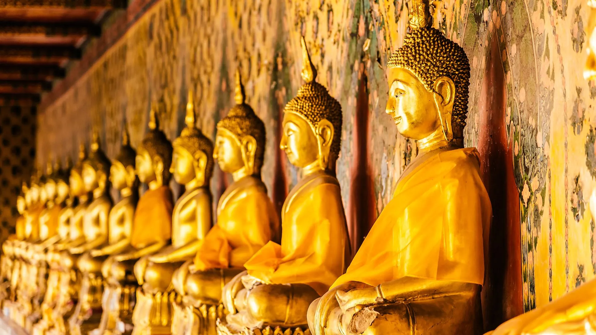 Fila de estátuas de Buda douradas e sentadas, uma ao lado da outra, dentro de um templo. As estátuas têm a mão direita pousada sobre a perna direita, na pose de meditação. A parede atrás delas é decorada com pinturas antigas.