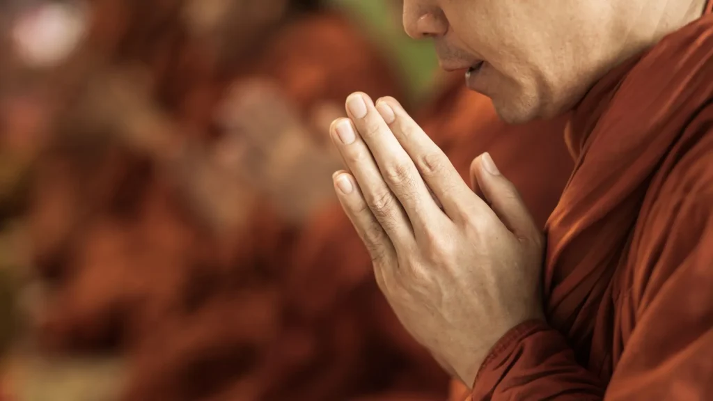 Mãos de um monge budista ou pessoa em meditação, com as palmas unidas em frente ao peito em sinal de respeito e oração. As mãos estão em foco, e a pessoa veste uma túnica cor de açafrão. O fundo está desfocado.