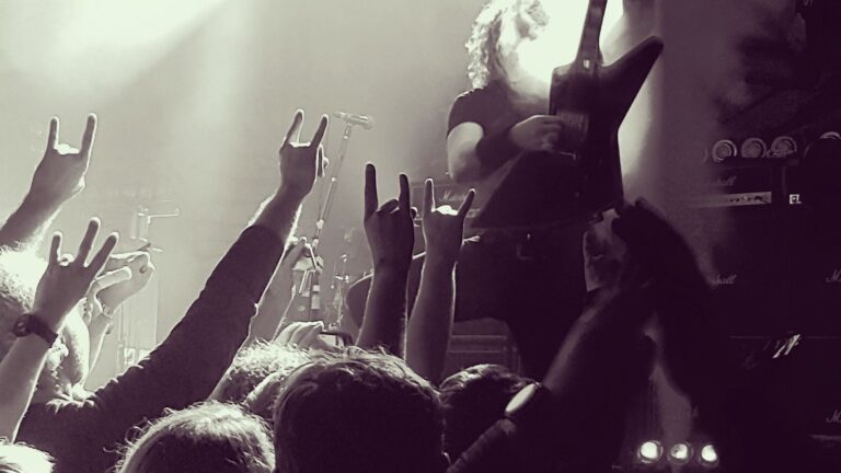 Multidão em um show de rock com os braços levantados, fazendo o gesto 'chifres do rock'. No centro, um guitarrista toca, iluminado por um holofote. A imagem é em preto e branco e a atmosfera é de energia e entusiasmo.