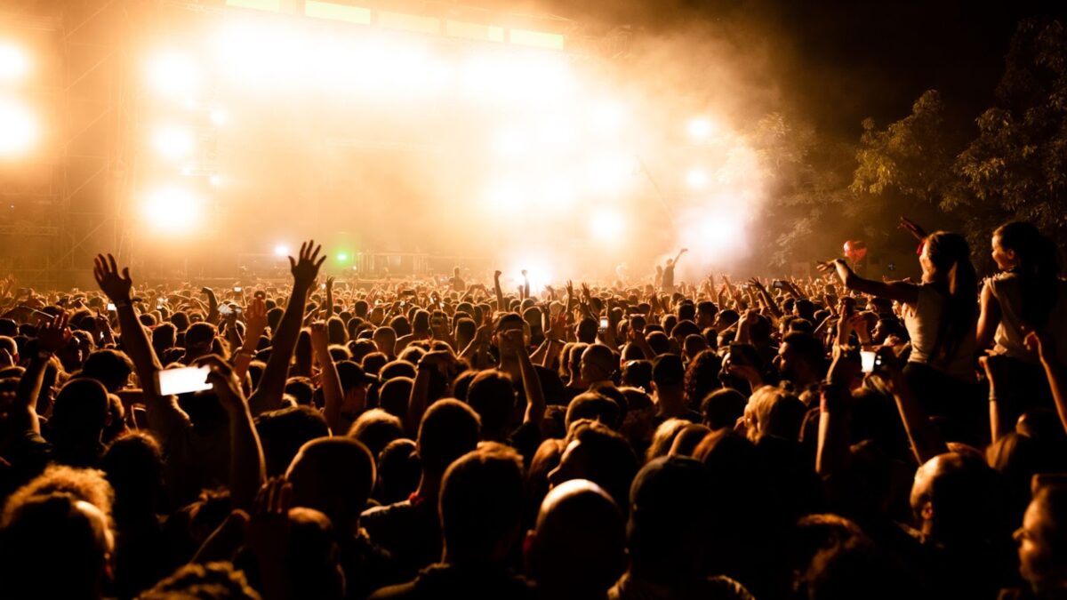 Grande multidão em um show ao ar livre à noite. As pessoas estão com os braços levantados e celulares capturando o momento. O palco está fortemente iluminado com luzes douradas e brancas, criando uma atmosfera festiva e energética.