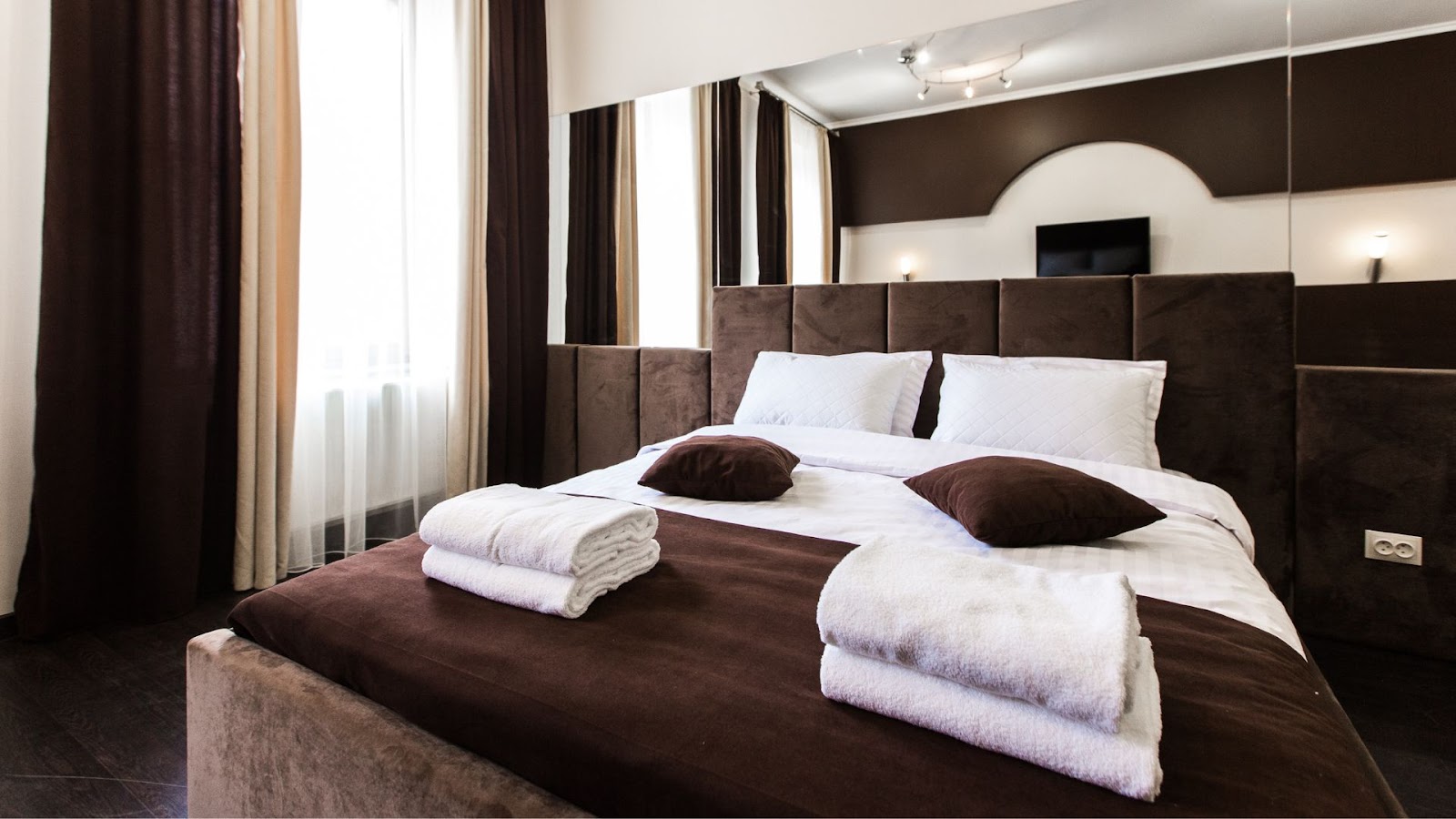 Uma cama arrumada em um quarto de hotel com lençóis brancos e travesseiros brancos e marrons. Duas toalhas brancas dobradas estão sobre a cama. As cortinas são de tons marrons e as paredes têm detalhes em marrom escuro, criando um ambiente aconchegante e luxuoso.