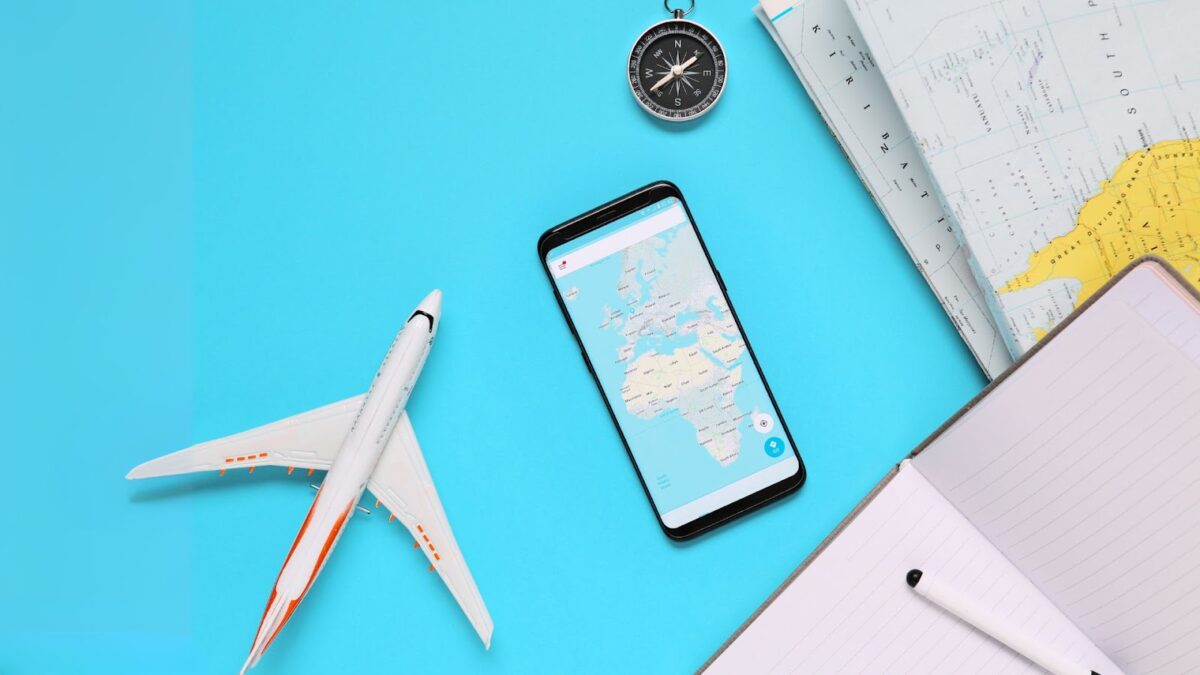 Objetos relacionados a viagens, como um pequeno avião de brinquedo, um smartphone mostrando um mapa, uma bússola e um caderno com uma caneta, dispostos sobre uma superfície azul clara. A imagem sugere planejamento ou preparação para uma viagem.