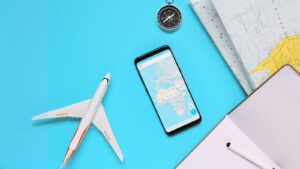 Objetos relacionados a viagens, como um pequeno avião de brinquedo, um smartphone mostrando um mapa, uma bússola e um caderno com uma caneta, dispostos sobre uma superfície azul clara. A imagem sugere planejamento ou preparação para uma viagem.