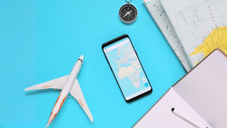 Objetos relacionados a viagens, como um pequeno avião de brinquedo, um smartphone mostrando um mapa, uma bússola e um caderno com uma caneta, dispostos sobre uma superfície azul clara. A imagem sugere planejamento ou preparação para uma viagem.