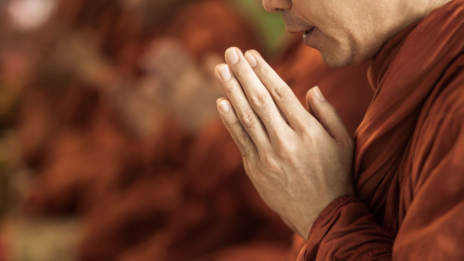 Mãos de um monge budista ou pessoa em meditação, com as palmas unidas em frente ao peito em sinal de respeito e oração. As mãos estão em foco, e a pessoa veste uma túnica cor de açafrão. O fundo está desfocado.
