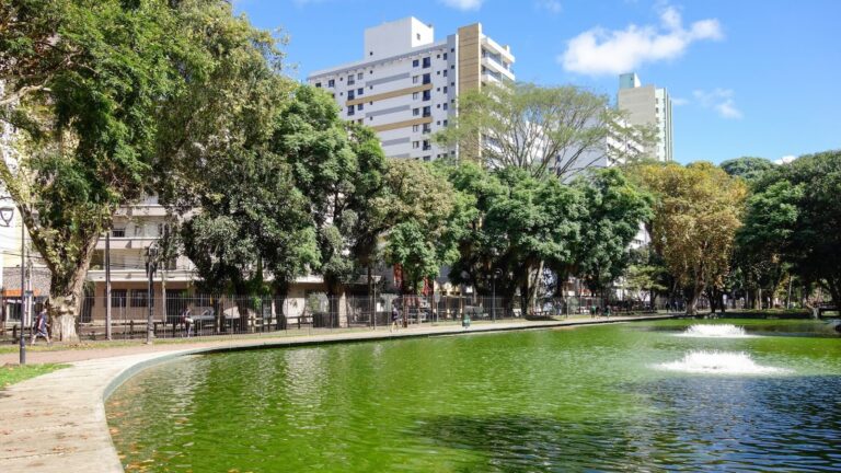 Vista do Passeio Público em Curitiba, Paraná, com um lago de água verde e uma fonte. A margem do lago é cercada por árvores frondosas, e prédios e edifícios altos podem ser vistos ao fundo, com um céu azul e poucas nuvens acima.