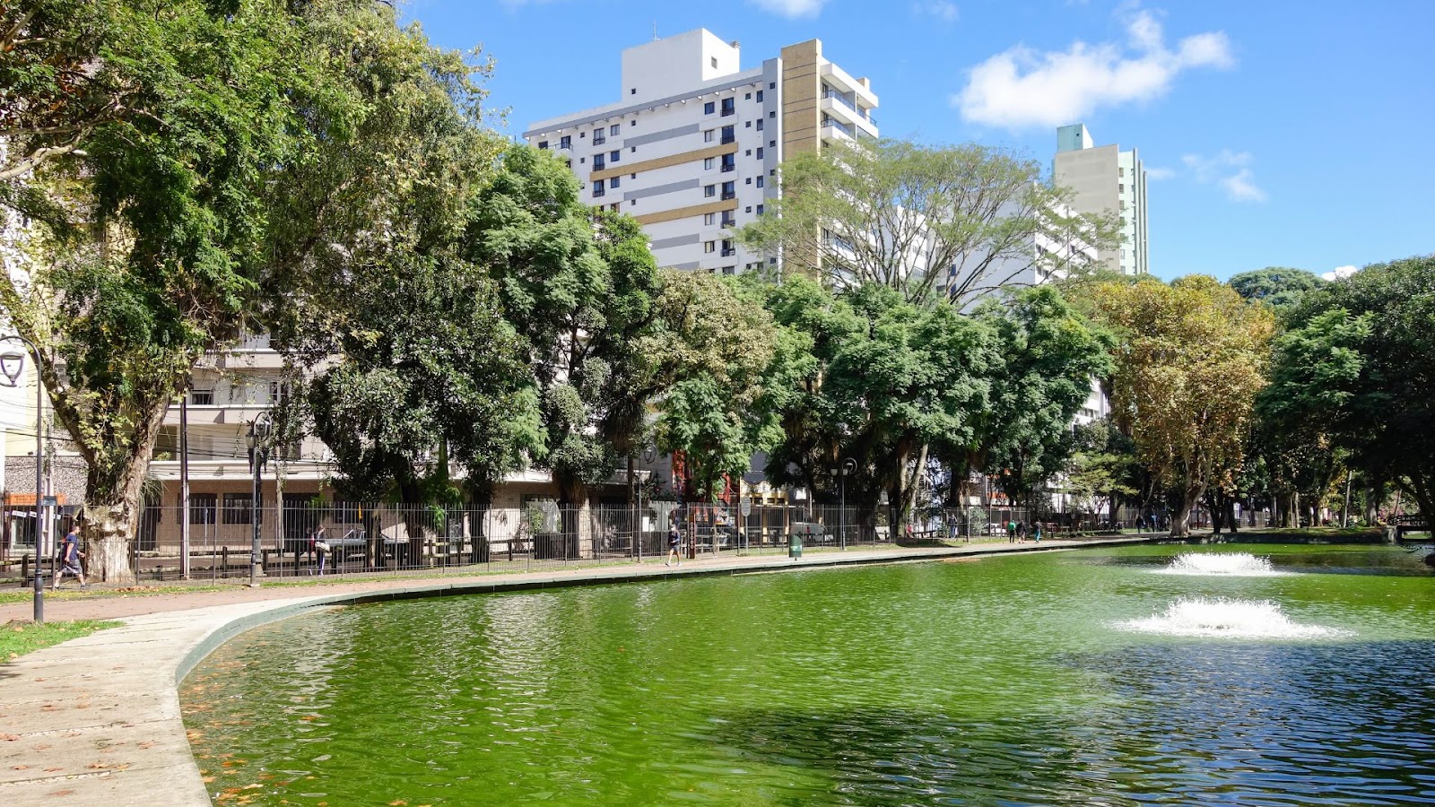 Vista do Passeio Público em Curitiba, Paraná, com um lago de água verde e uma fonte. A margem do lago é cercada por árvores frondosas, e prédios e edifícios altos podem ser vistos ao fundo, com um céu azul e poucas nuvens acima.
