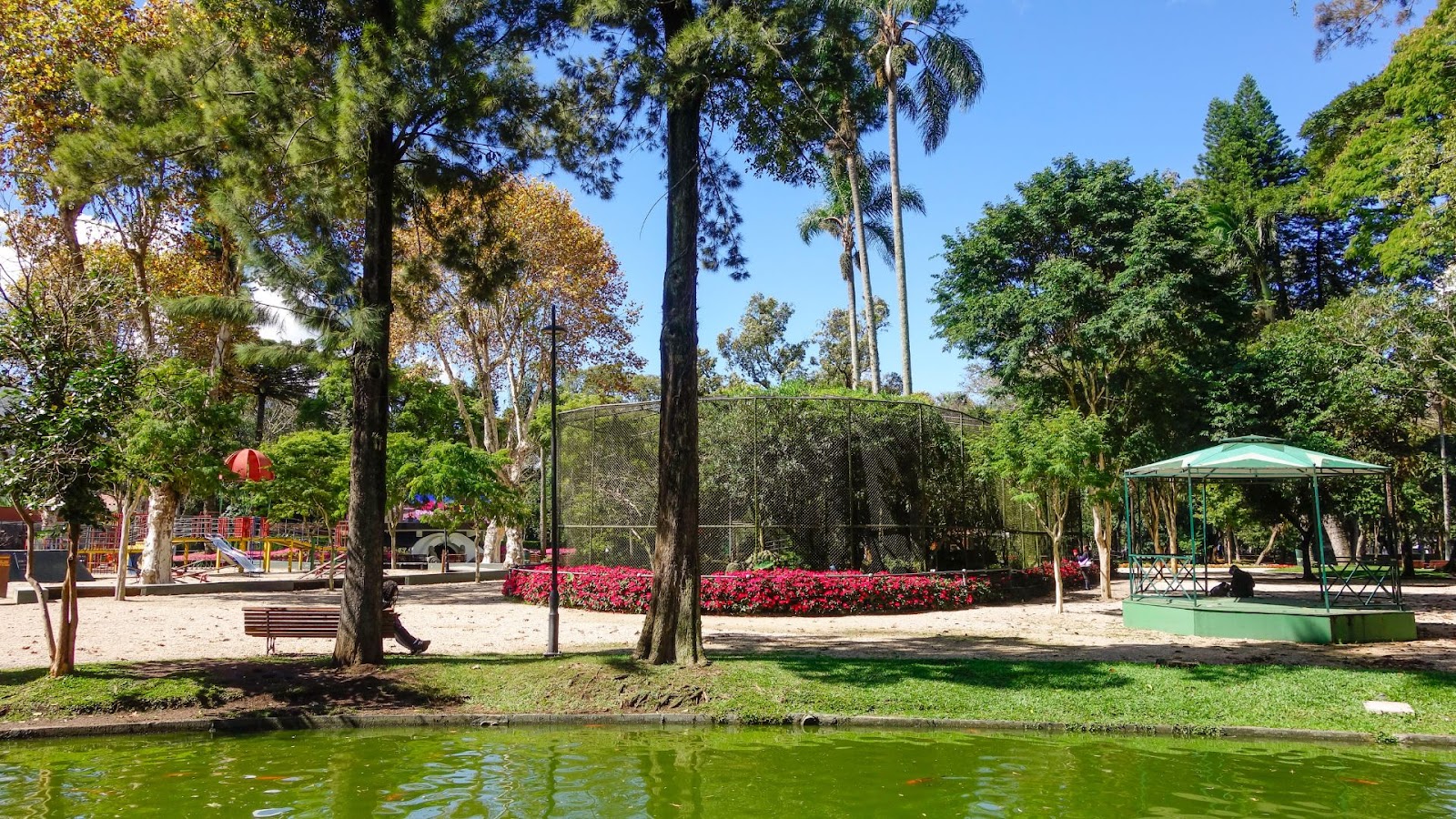 Vista de uma área do Passeio Público de Curitiba, no Paraná. Em primeiro plano, um lago de água verde. Ao centro, um grande viveiro de pássaros cercado por flores vermelhas. Do lado direito, um coreto verde com telhado. Do lado esquerdo, um parquinho infantil com balanços. A cena é cercada por diversas árvores.