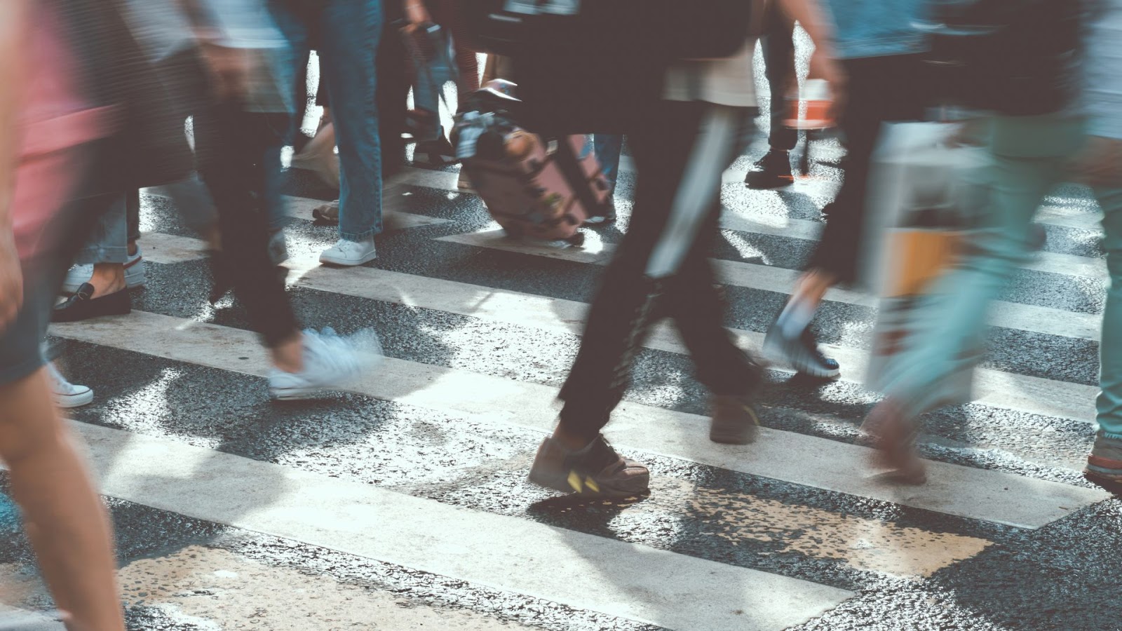 Multidão de pessoas cruzando a rua em uma faixa de pedestres. As pessoas estão em movimento, criando um efeito de desfoque, o que indica velocidade. O foco está nos pés e pernas das pessoas, e as linhas brancas e pretas da faixa de pedestres são visíveis.