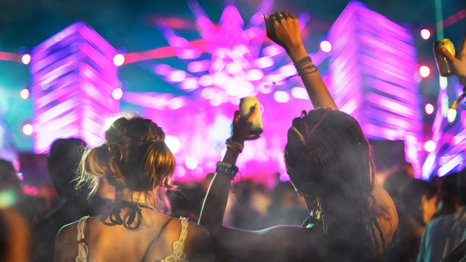 Duas pessoas de costas, com os braços levantados, assistindo a um show ao ar livre à noite. O palco está iluminado com luzes roxas e coloridas, criando um ambiente animado de festival.