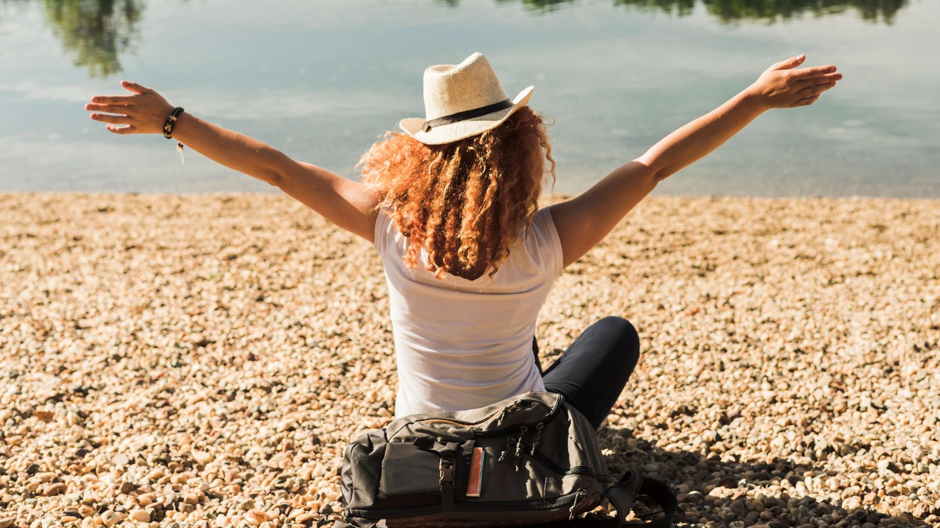 Uma mulher de chapéu de palha e cabelo ruivo e cacheado, sentada de costas para a câmera, de frente para um lago. Ela está com os braços abertos, em um gesto que sugere liberdade ou felicidade. Ela está sentada na margem de um lago, em uma área coberta por pequenas pedras e areia. A mulher usa uma blusa branca e calça escura, e há uma mochila escura na frente dela.