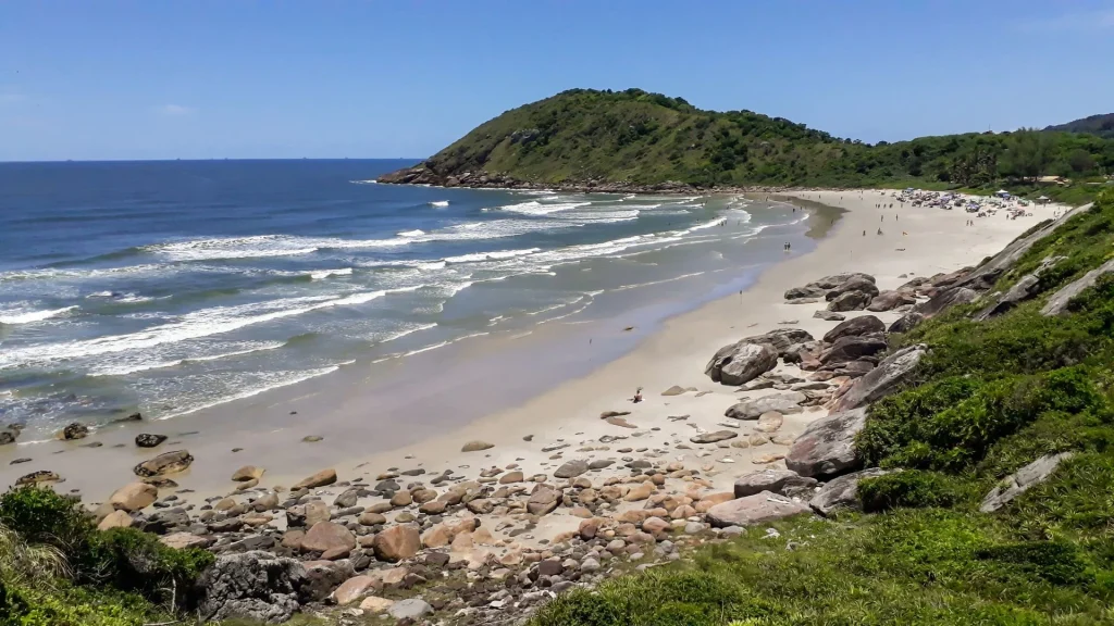 Uma vista panorâmica de uma praia de areia dourada e mar azul, com ondas quebrando suavemente. Há uma grande colina verde e rochosa no lado direito da praia, e algumas pessoas podem ser vistas tomando sol na areia. Grandes pedras e rochas, algumas com musgo, cobrem a encosta em primeiro plano. O céu está limpo e azul.