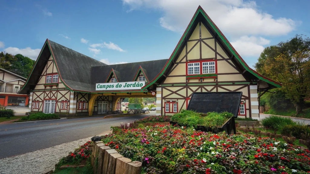 Uma fotografia diurna do portal de entrada de Campos do Jordão, em estilo arquitetônico suíço-alpino. A estrutura tem telhados íngremes e é decorada com detalhes em madeira escura sobre paredes claras. Há uma placa verde com o nome "Campos do Jordão" no arco central. Na frente do portal, há canteiros de flores coloridas e grama, e a imagem é capturada em um dia com céu parcialmente nublado.