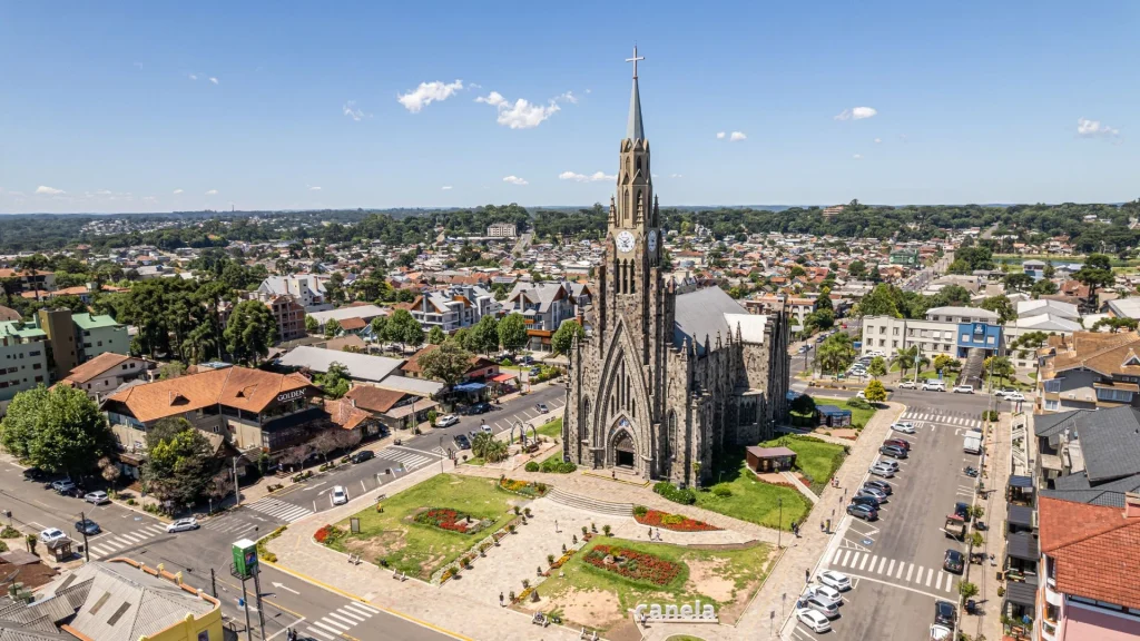Uma visão aérea de uma cidade, mostrando uma majestosa igreja de estilo gótico com uma torre alta e pontiaguda no centro. A igreja, feita de pedra escura, está em frente a uma praça verde com canteiros de flores. A cidade ao redor é composta por prédios de telhados vermelhos e outras construções, cercadas por muitas árvores. O céu está azul e com poucas nuvens, indicando um dia ensolarado.
