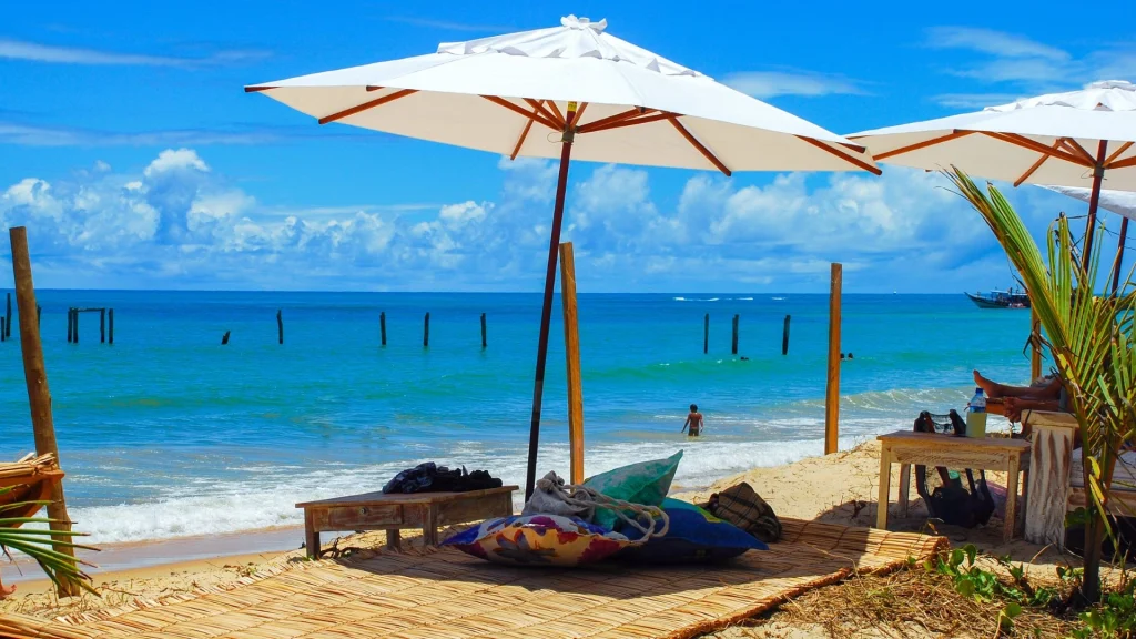 Uma cena de praia tropical em um dia ensolarado. A imagem mostra um espaço de descanso com esteiras de palha no chão e almofadas coloridas, à sombra de grandes guarda-sóis brancos. Há uma espreguiçadeira de madeira e uma mesa na areia. O mar azul-turquesa, com pequenas ondas, se estende até o horizonte, onde barcos flutuam. No mar, há estacas de madeira e uma pessoa tomando banho. O céu é azul e com algumas nuvens brancas.