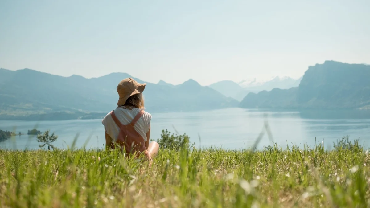 Imagem de uma pessoa sentada em um campo de grama verde, de costas para a câmera, olhando para um grande lago cercado por montanhas ao fundo. A pessoa usa um chapéu de tecido, uma roupa clara e mochila de cor salmão. O céu está limpo e azul, e a cena transmite sensação de tranquilidade e contato com a natureza.