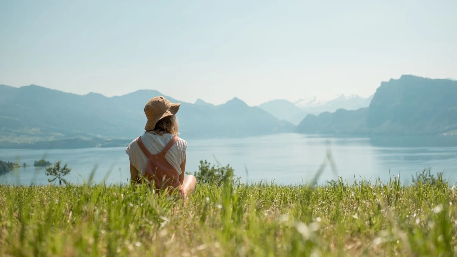 Imagem de uma pessoa sentada em um campo de grama verde, de costas para a câmera, olhando para um grande lago cercado por montanhas ao fundo. A pessoa usa um chapéu de tecido, uma roupa clara e mochila de cor salmão. O céu está limpo e azul, e a cena transmite sensação de tranquilidade e contato com a natureza.
