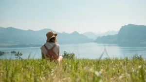 Imagem de uma pessoa sentada em um campo de grama verde, de costas para a câmera, olhando para um grande lago cercado por montanhas ao fundo. A pessoa usa um chapéu de tecido, uma roupa clara e mochila de cor salmão. O céu está limpo e azul, e a cena transmite sensação de tranquilidade e contato com a natureza.