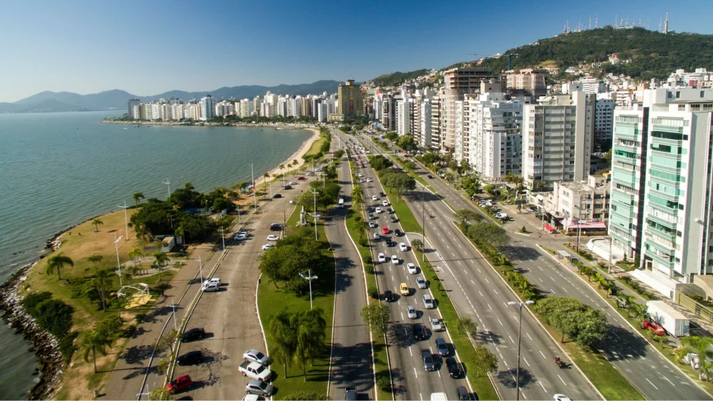 Vista aérea da Beira-Mar Norte, em Florianópolis. A imagem mostra a movimentada avenida com carros, o mar calmo do lado esquerdo e uma orla cheia de árvores e palmeiras. Ao fundo, uma cidade com vários prédios altos e morros. O céu é azul e sem nuvens.
